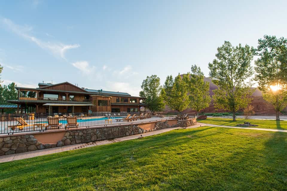 Rustic wooden lodge with large pool, stone features, manicured lawns, and evening sunset lighting