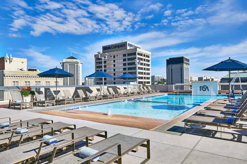 Urban rooftop pool with blue umbrellas, loungers, cityscape skyline, and downtown buildings visible