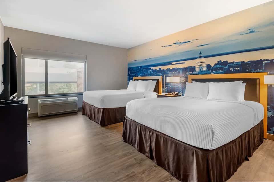 Hotel guest room with two queen beds, cityscape mural headboard, and window with landscape view