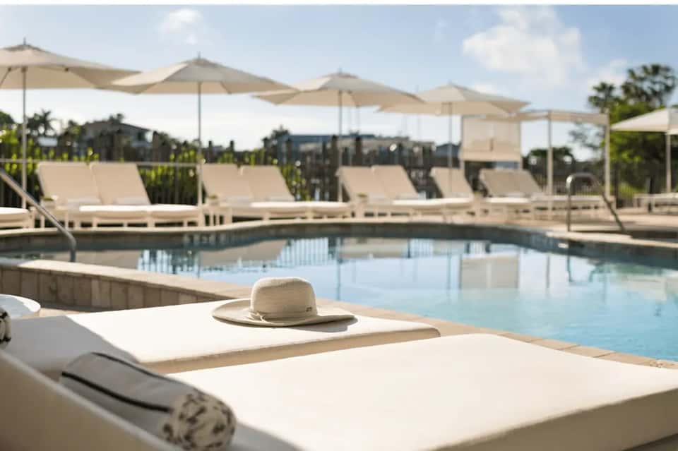Resort pool area with white lounge chairs, tan umbrellas, cypresses, and peaceful water reflection