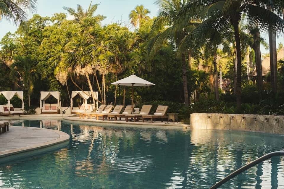 Resort pool area with white cabanas, lounge chairs, palm trees, and manicured landscaping