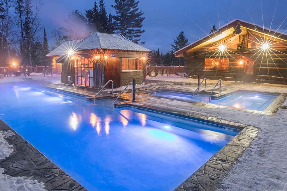 Illuminated hot springs pool complex at dusk with wooden pavilions, glowing blue water, snow, and forest surroundings