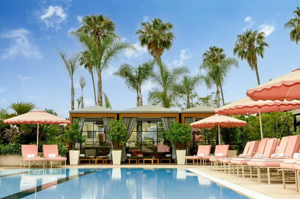 Resort pool with pink lounge chairs, modern cabanas, and palm trees under blue sky