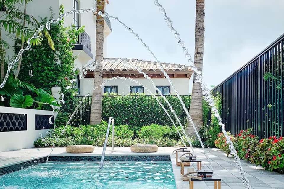 Intimate courtyard pool with modern lounge chairs, white palm trees, waterfall feature, and tropical greenery