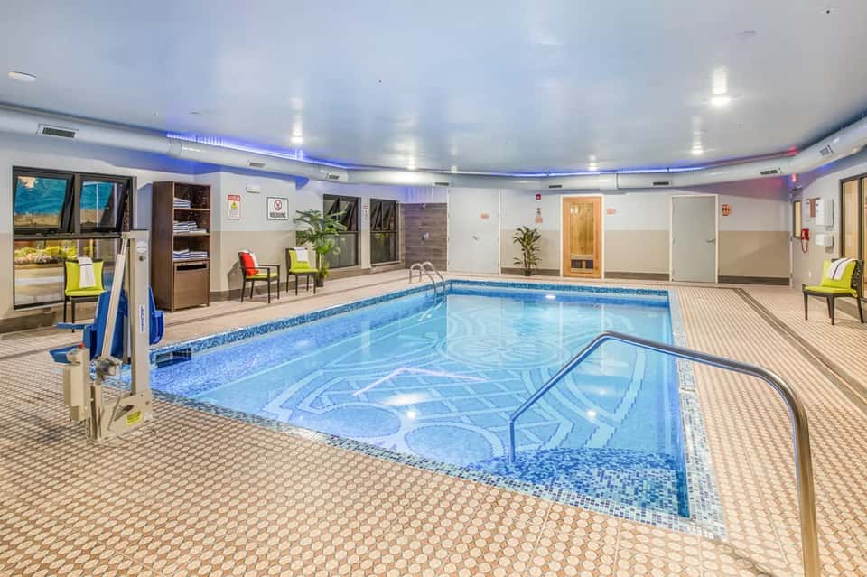 Indoor heated swimming pool with blue water, deck chairs, glass doors, and modern amenities