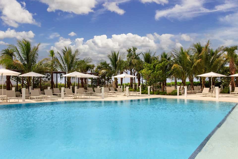 Resort pool surrounded by white umbrellas, loungers, palm trees, and white cabanas under blue sky with clouds