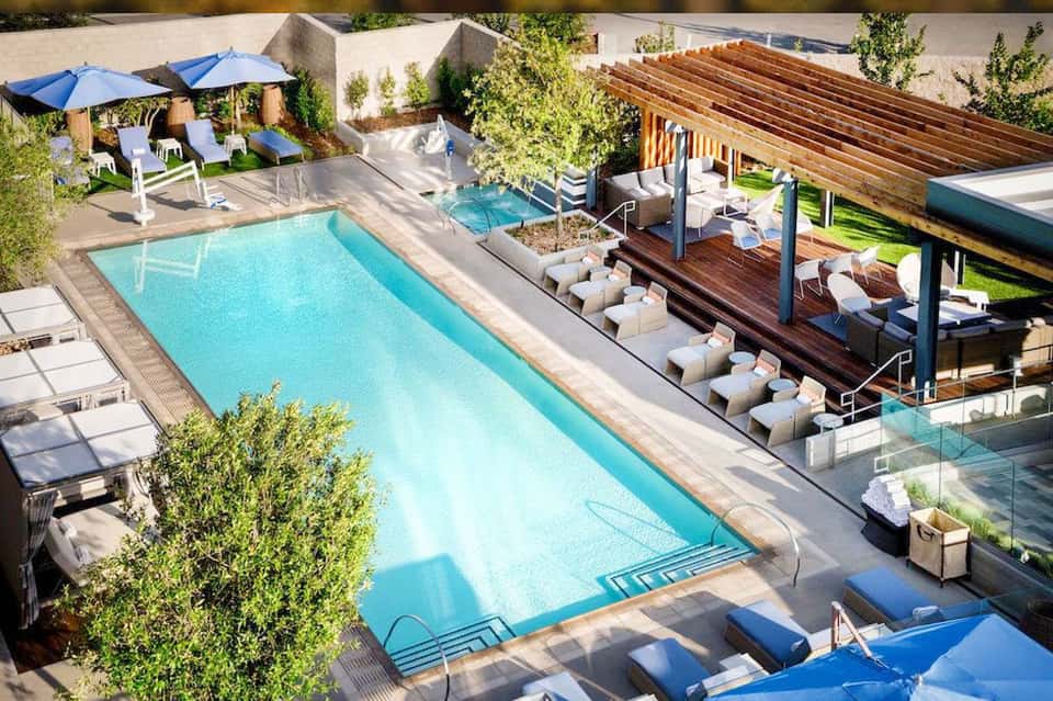 Aerial view of resort pool with wooden cabanas, loungers, and surrounding landscaping