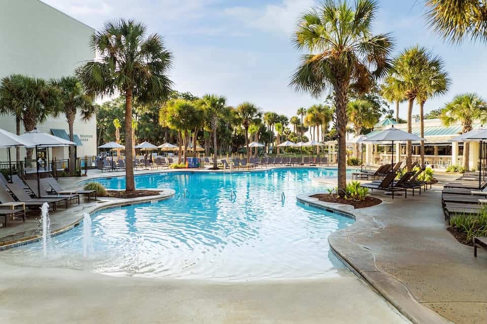 Resort pool surrounded by palm trees with lounge chairs, umbrellas, and tropical landscaping