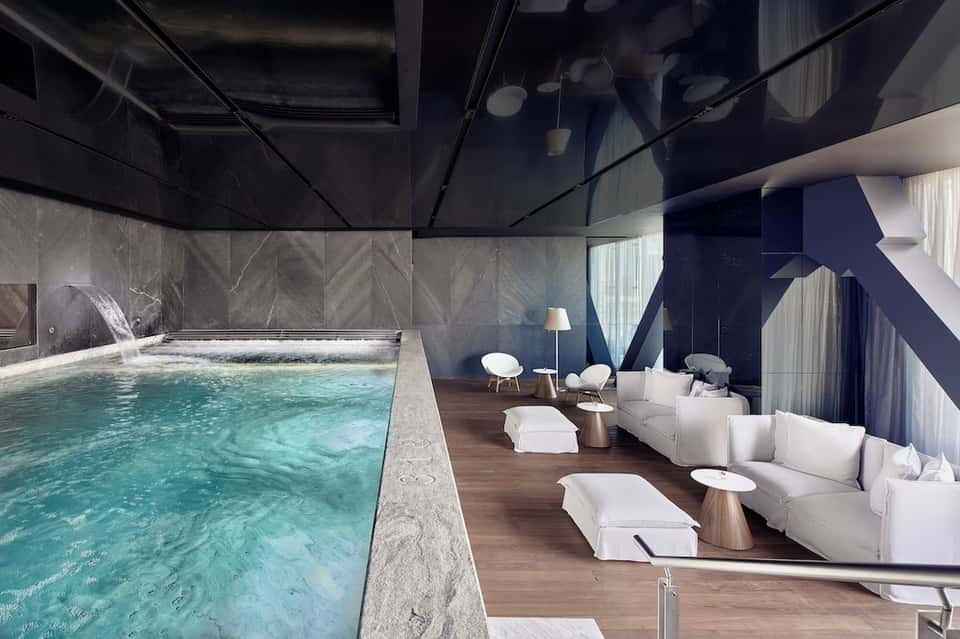 Luxurious indoor heated pool with marble surround, floor-to-ceiling windows, white seating, and modern architectural design