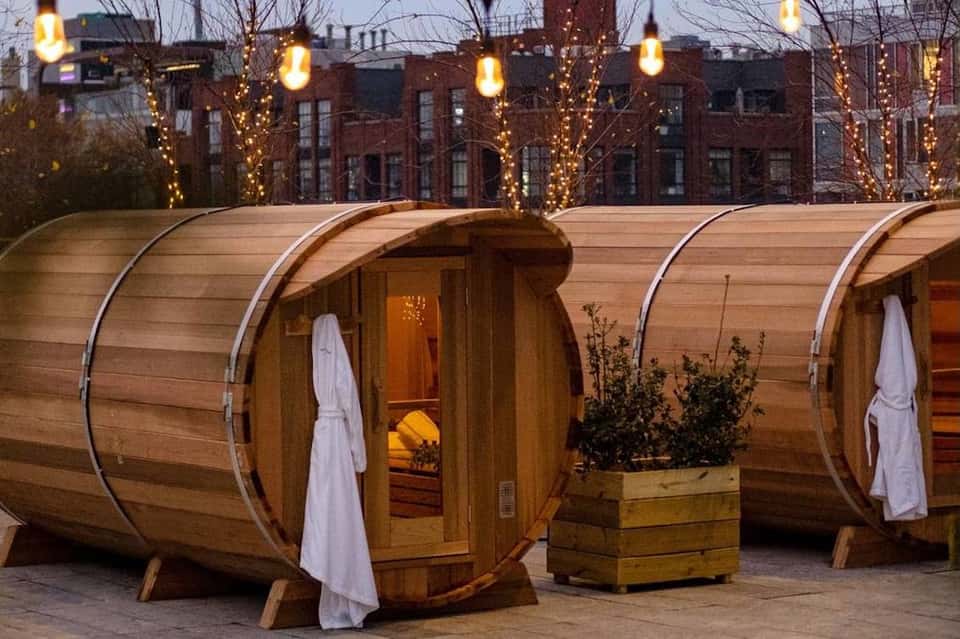Wooden barrel-shaped spa pods with white curtains lit by hanging lanterns in urban courtyard setting