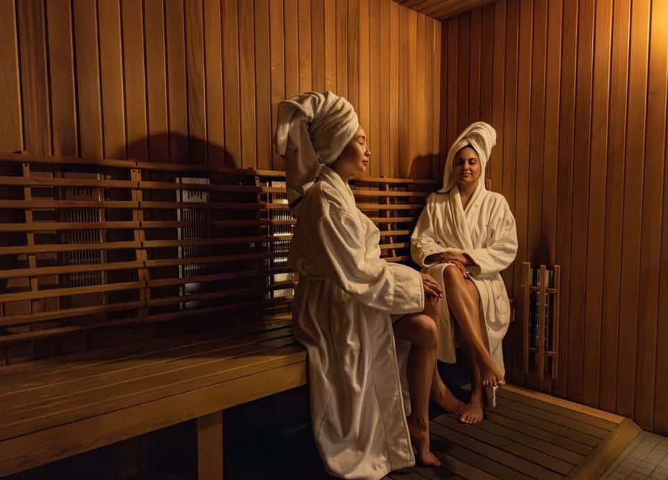 Two women in spa robes relaxing in a sauna with wooden benches and warm lighting