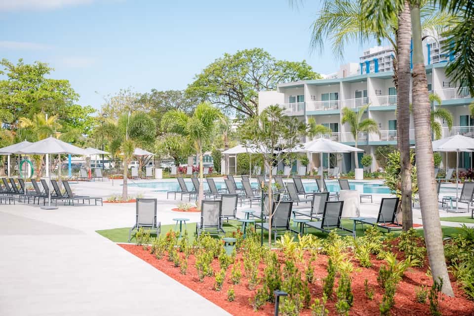 Resort pool area with lounge chairs, umbrellas, palm trees, and landscaped grounds