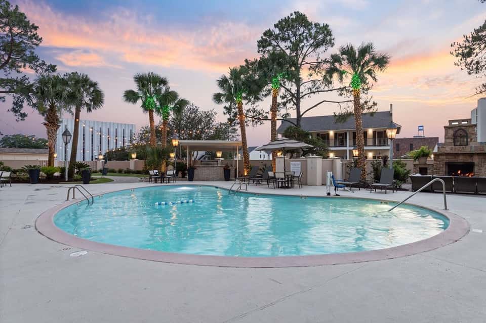 Resort pool at dusk with palm trees, lounge chairs, fireplace, and lit gazebo
