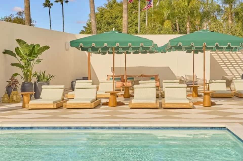 Pool area with green umbrellas over wooden lounge chairs, concrete deck, palm trees, and beige walls