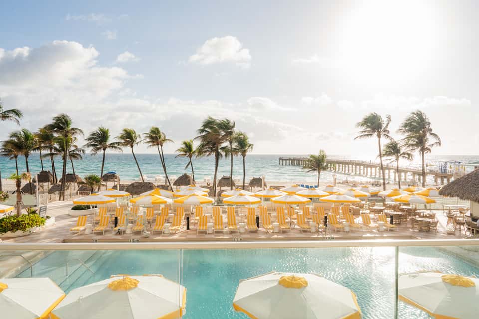 Beachfront resort pool terrace with yellow umbrellas, palm trees, ocean pier, and ocean views
