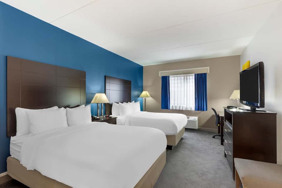 Hotel guest room with two queen beds, blue accent wall, brown headboards, desk area, and large window with blue curtains