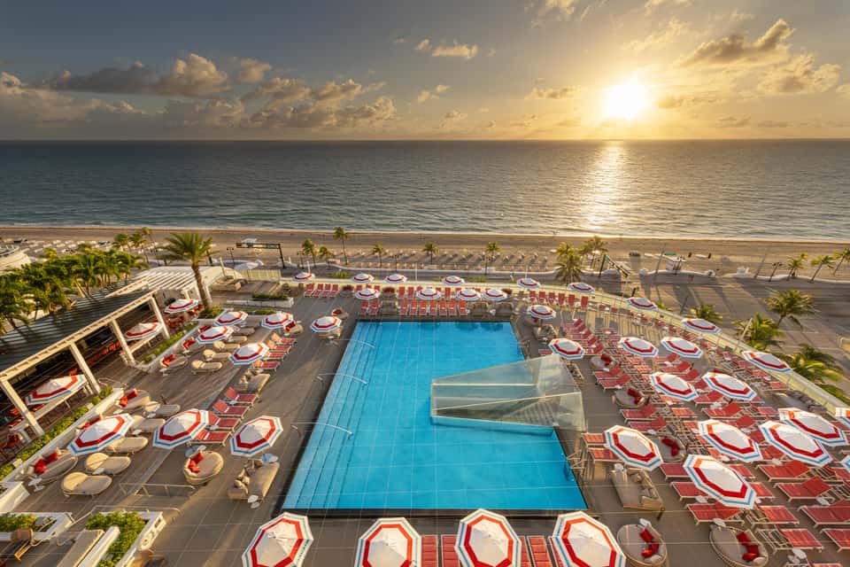Aerial view of oceanfront resort pool at sunset with red umbrellas and beach