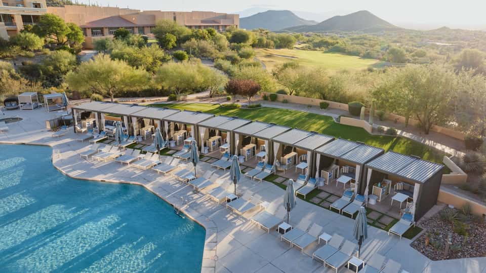 Resort pool with turquoise water, lounge chairs, cabanas, mountain valley views, and olive grove landscape