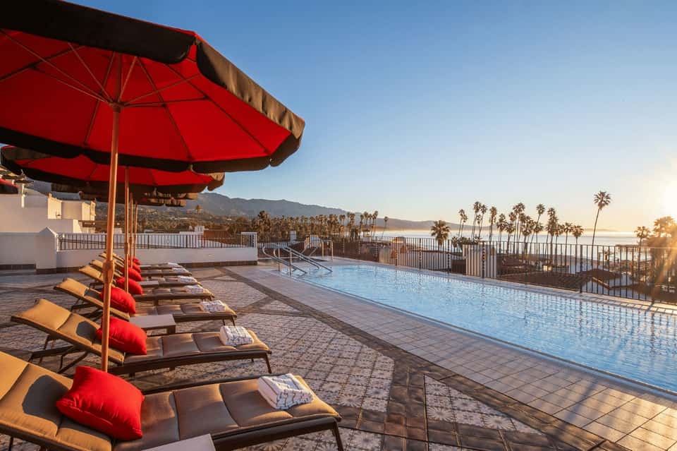 Hilltop pool deck at sunset with red umbrellas, lounge chairs, and panoramic mountain and ocean views