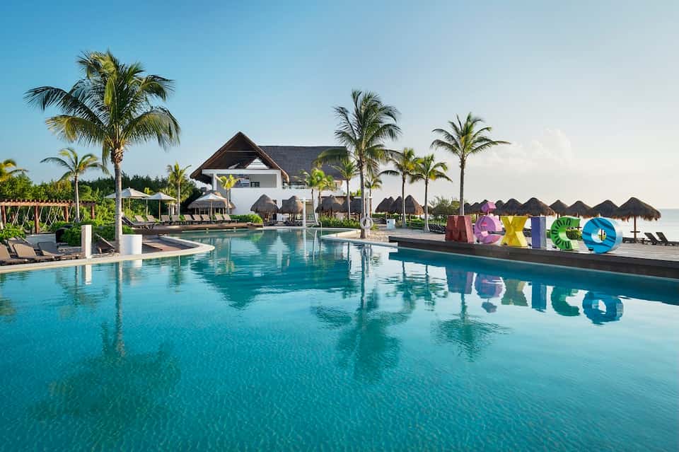 Modern resort pool with turquoise water, white buildings, palm trees, and colorful letters spelling resort name