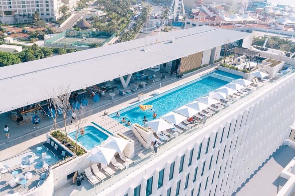 Aerial view of modern resort with rectangular pools, lounge chairs, and city surroundings