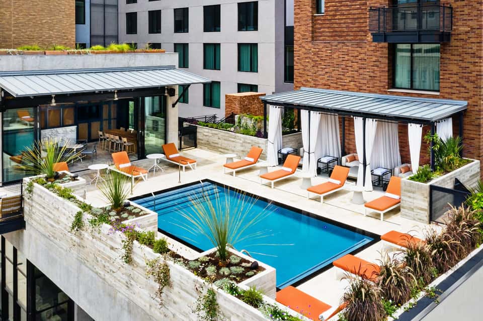 Aerial view of rooftop pool with orange loungers, white cabanas, and surrounding brick buildings
