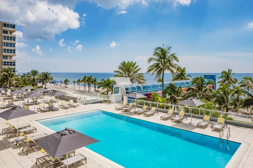 Oceanfront resort pool with lounge chairs, palm trees, and beachfront views under blue sky