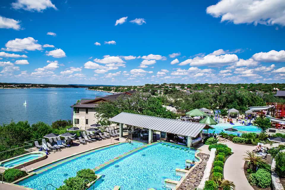 Aerial view of lakeside resort with turquoise swimming pools, lounge chairs, and water views under blue sky