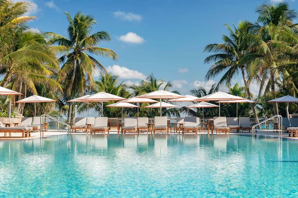 Luxury resort pool with turquoise water, white umbrellas, wooden loungers, and palm trees under blue sky
