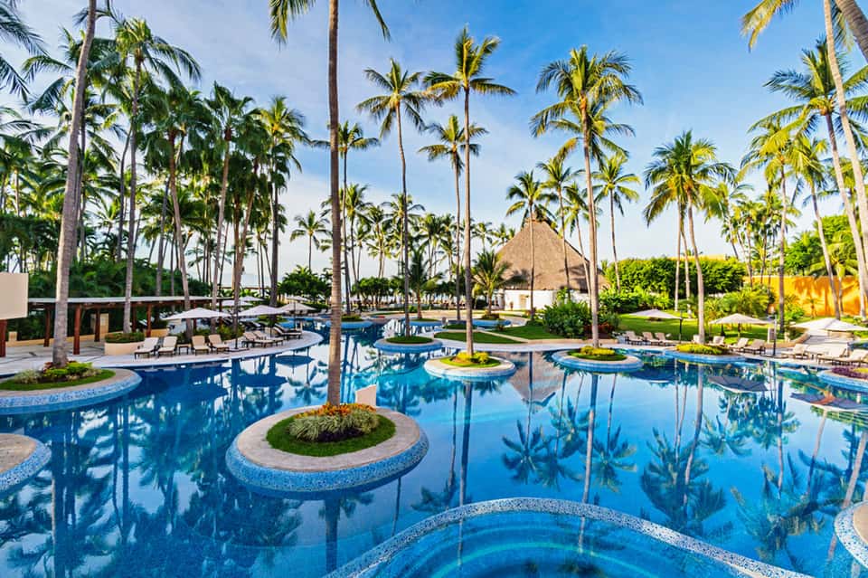 Luxury resort pool complex with crystal-blue water, palm trees, thatched-roof structures, and manicured lawns