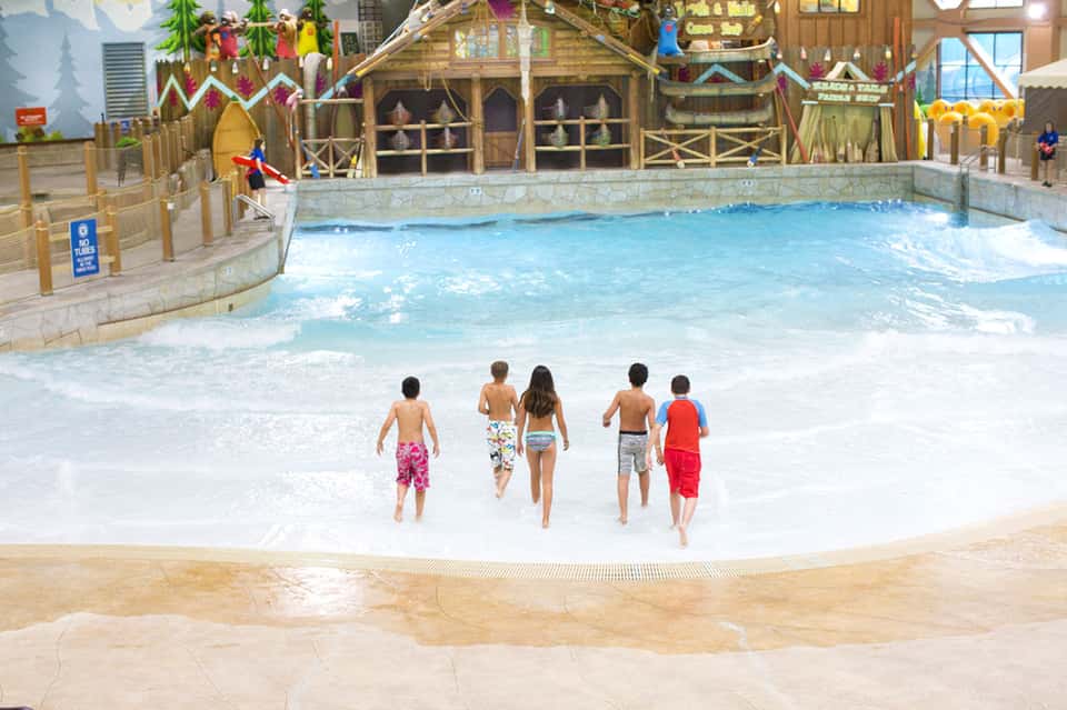 Children wading into indoor wave pool with tropical resort theming and thatched structures
