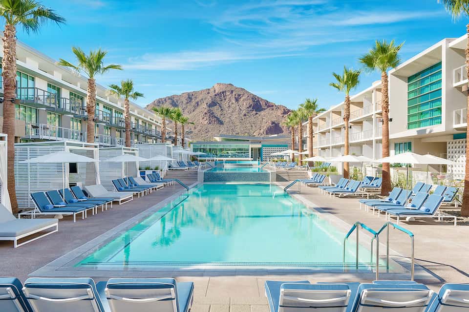 Resort pool with turquoise water, palm trees, modern white buildings, and desert mountain backdrop
