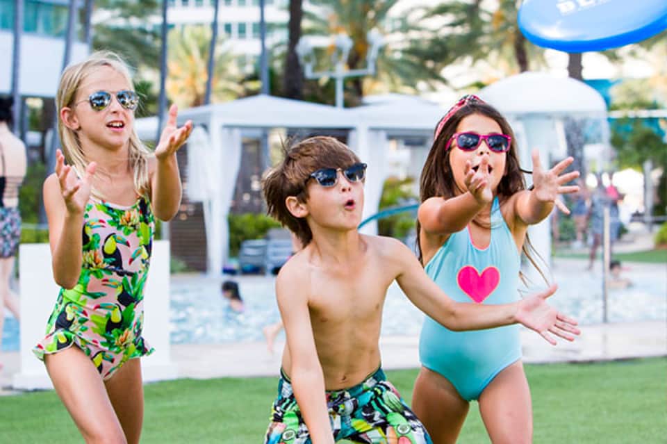Three children in colorful swimsuits playing on grass near a resort pool area