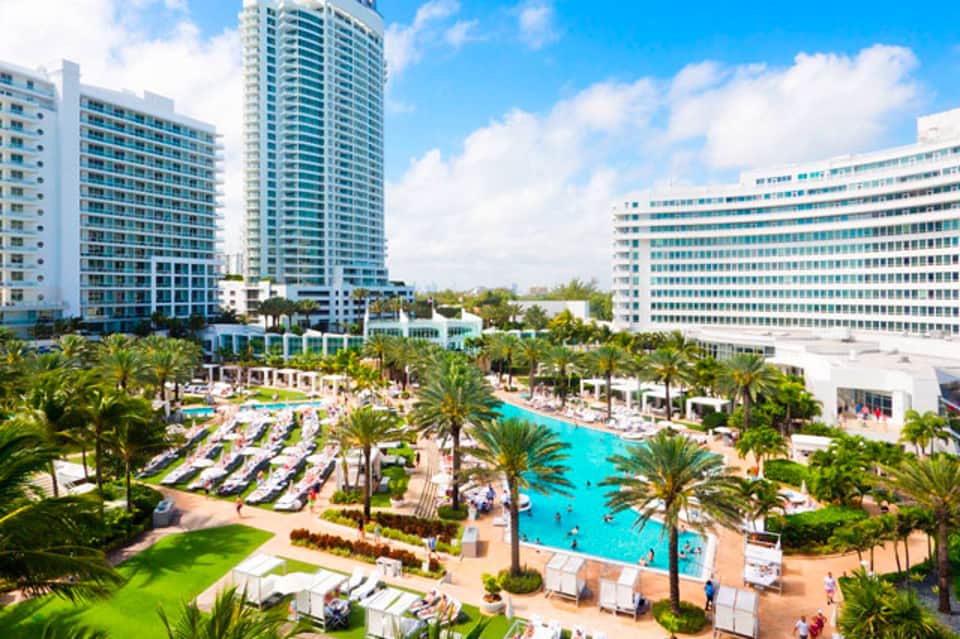Aerial view of resort with turquoise pool, palm trees, lounge chairs, and multi-story hotel towers under blue sky