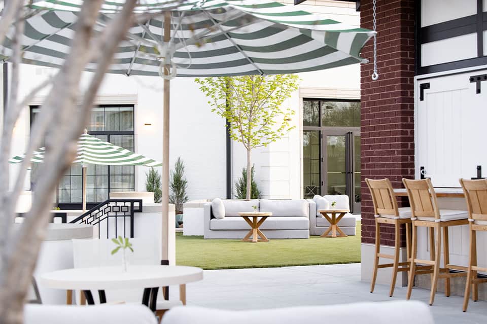 Modern courtyard with striped umbrella, lounge seating, wooden chairs, and manicured lawn surrounded by brick and white buildings