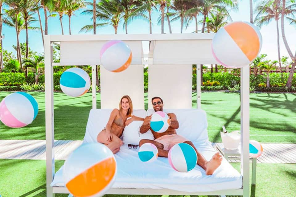 Couple relaxing on a bed under a white canopy with colorful beach balls in a tropical garden setting