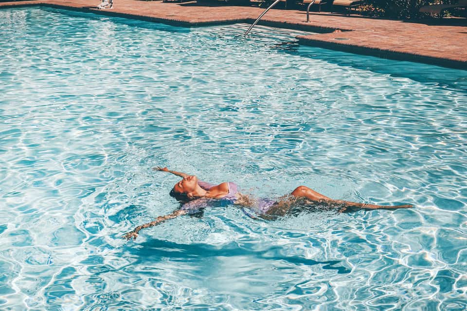 Woman floating peacefully in crystal-clear turquoise pool water with wooden dock and green landscape
