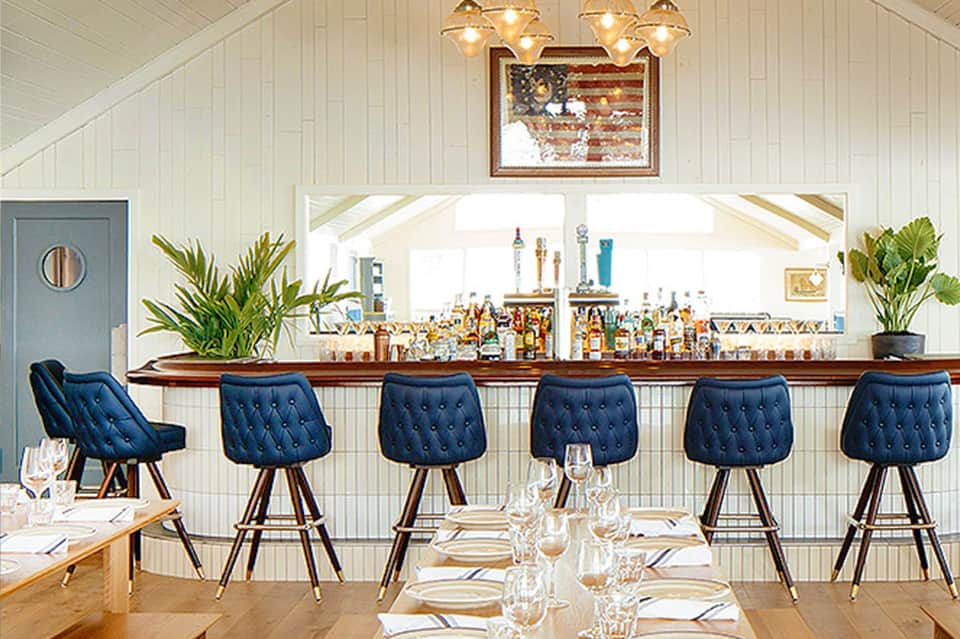 Elegant dining room with navy tufted bar stools, wooden bar counter, white shiplap walls, and brass chandelier lighting