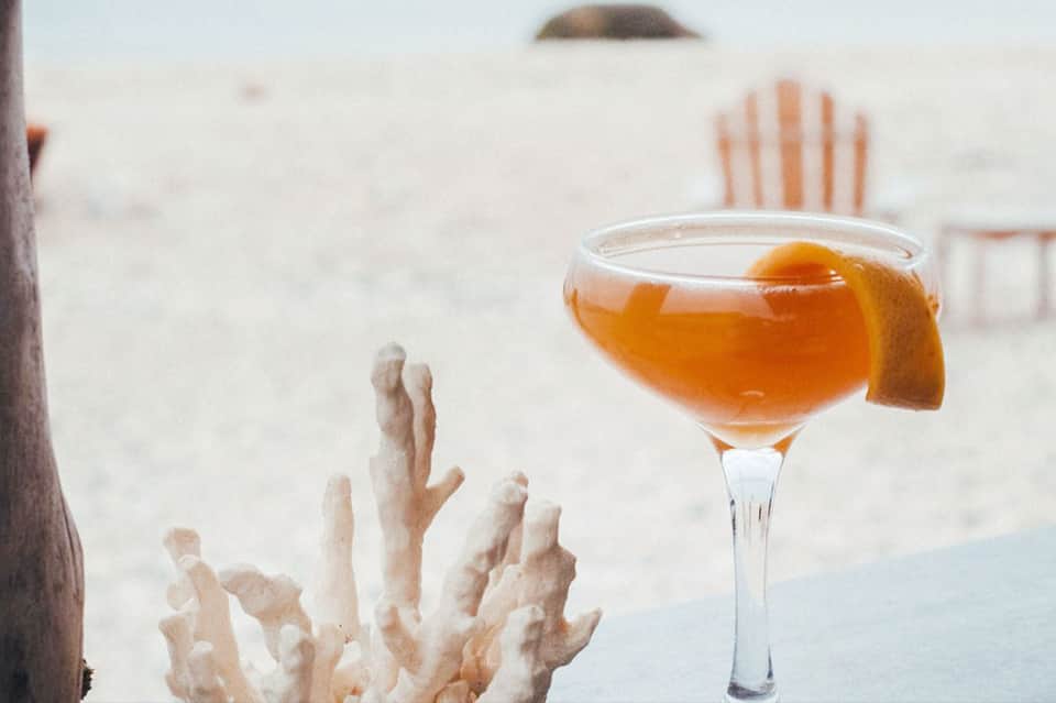 Amber cocktail with orange slice garnish on beach with coral backdrop