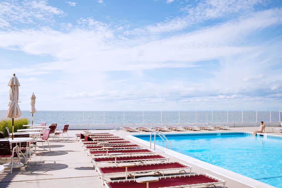 Outdoor pool deck with burgundy lounge chairs, umbrellas, and ocean view behind white fence