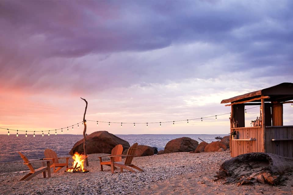 Bonfire on beach at dusk with Adirondack chairs, wooden structure, and string lights over ocean