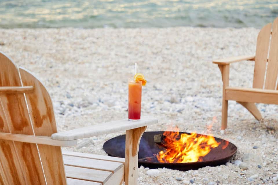 Beach scene with Adirondack chairs, fire pit, cocktail, and ocean waves at sunset