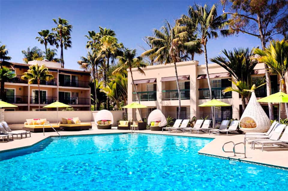 Resort pool with turquoise water surrounded by palm trees, lounge chairs, and colorful umbrellas