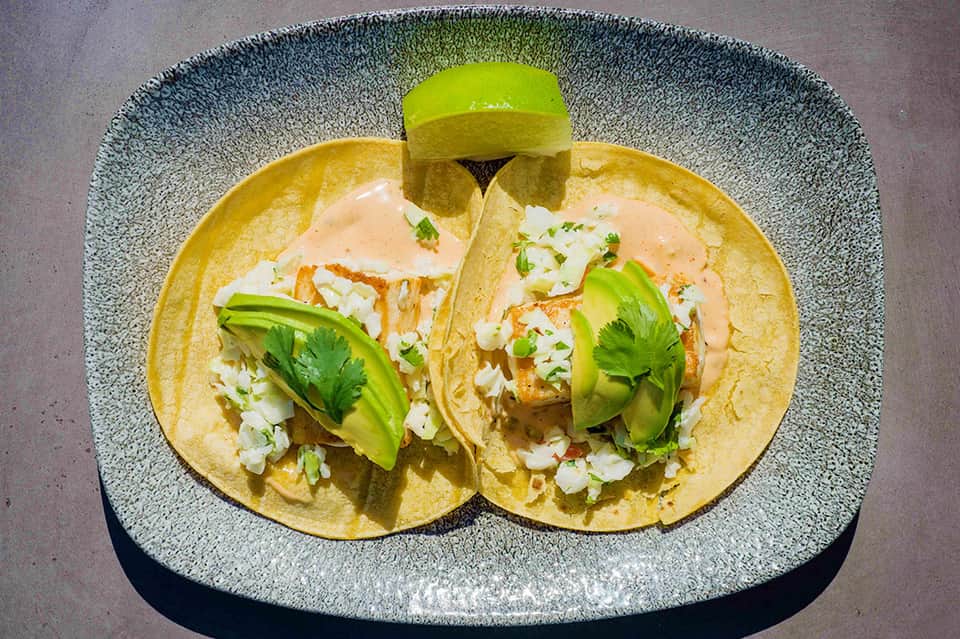 Two fish tacos with avocado, cilantro, and white onions on a textured plate