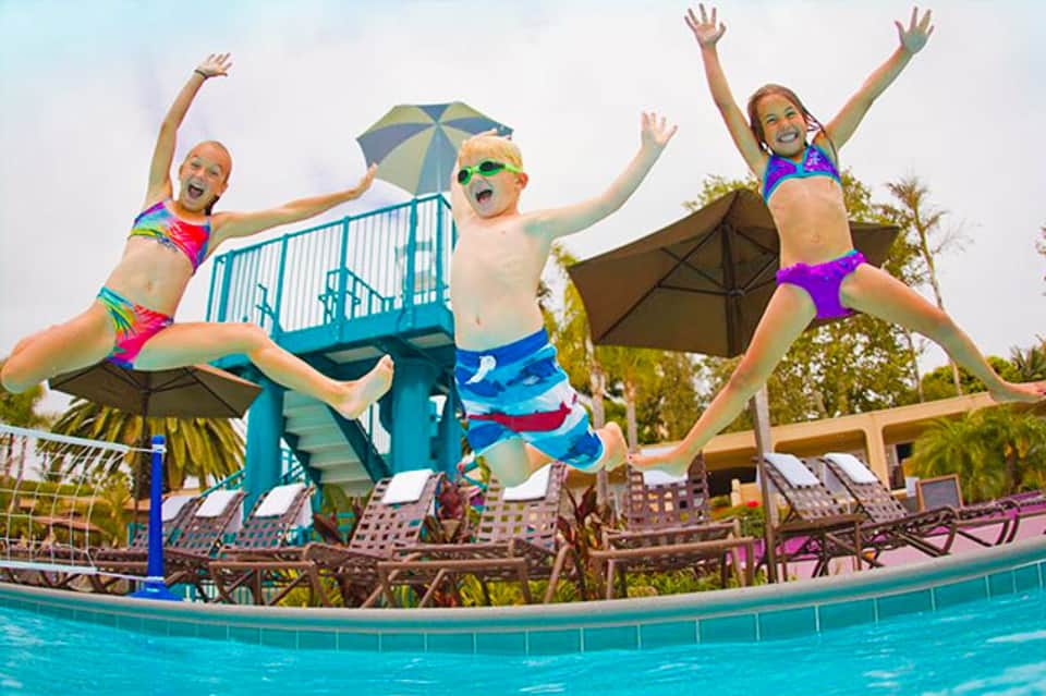 Three children jumping into a swimming pool with umbrellas and loungers in background