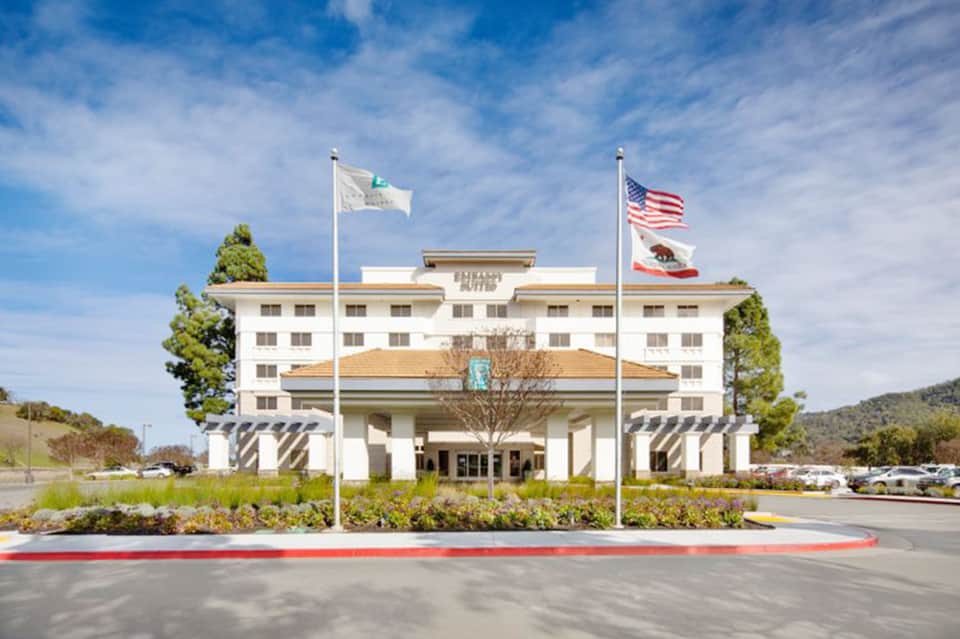 Embassy Suites hotel exterior with white facade and flags flying in front of landscaped grounds