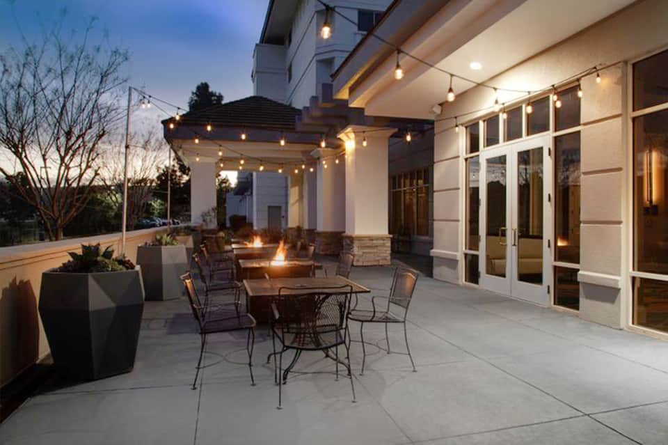 Modern patio with fire pit, wooden pergola, string lights, and contemporary furnishings at dusk
