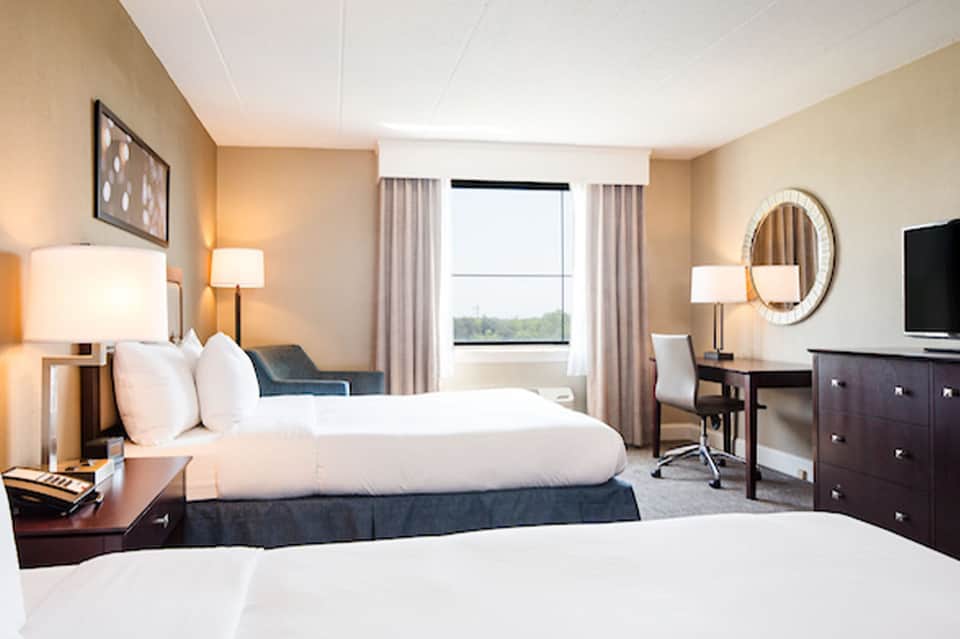 Modern hotel bedroom with two queen beds, work desk, and large windows overlooking green landscape