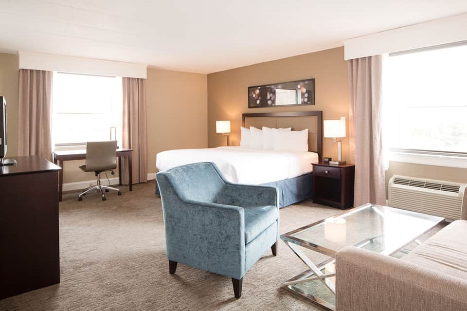 Modern hotel bedroom with queen bed, blue accent chair, work desk, and warm earth-tone décor