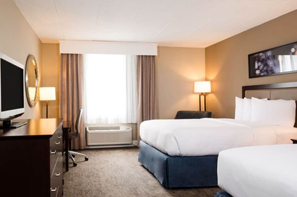 Hotel room with two queen beds, warm neutral tones, work desk, TV, and window with curtains.